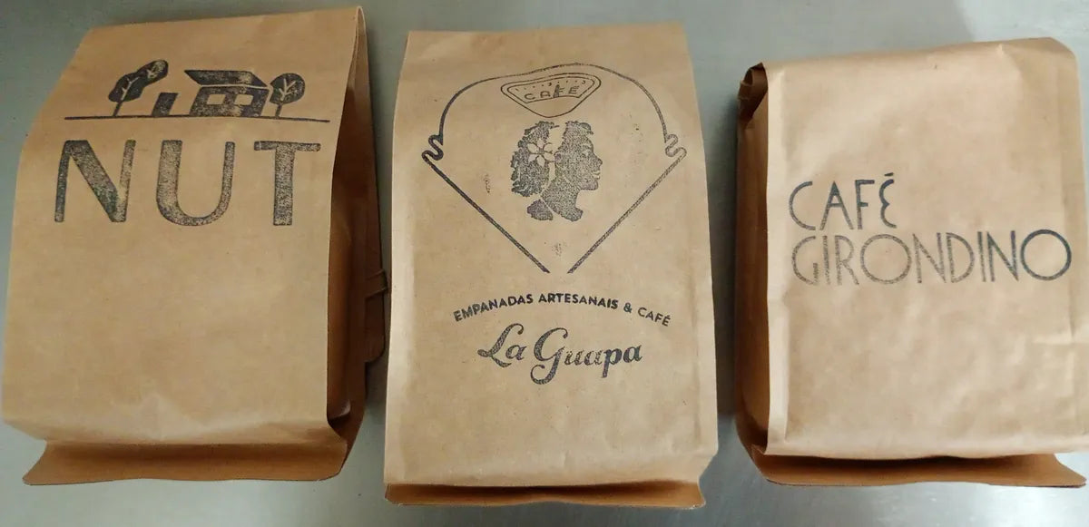 Três pacotes kraft de café carimbados com as marcas NUT, La Guapa e Café Girondino